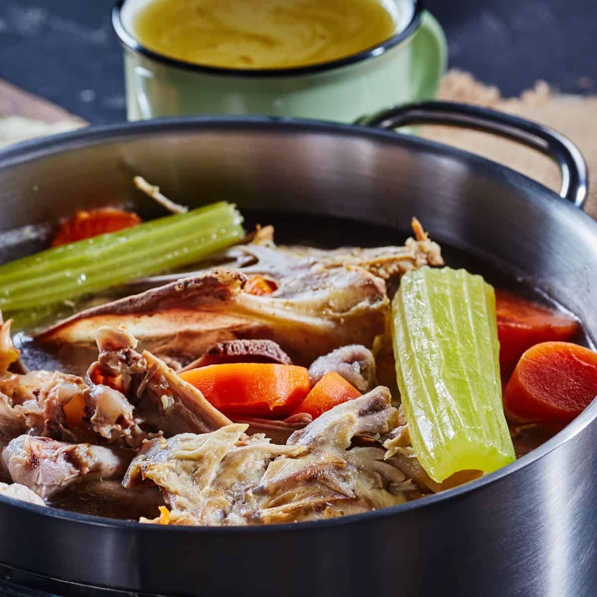 Metal cooking pot with celery stalks, carrot slices and chicken carcass in water.