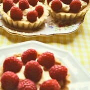 Three small tarts with a cream filling topped with red fruit on a white plates, with a yellow gingham table cloth.