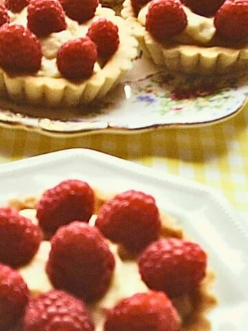 Three small tarts with a cream filling topped with red fruit on a white plates, with a yellow gingham table cloth.