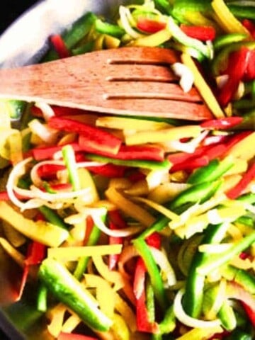 Sliced green, red, and yellow peppers filling a sauteed pan with a slotted wood spoon.