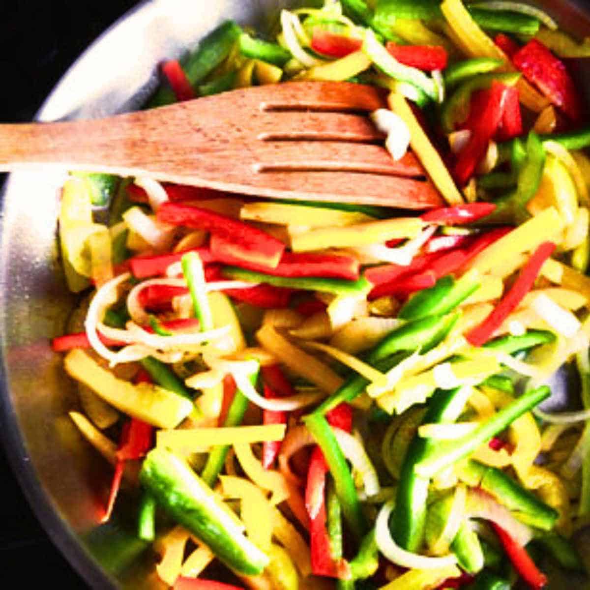 Sliced green, red, and yellow peppers filling a sauteed pan with a slotted wood spoon.