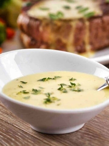 White bowl with cream colored sauce sprinkled with green leaves, a spoon sitting in bowl and a thick piece of beef with sauce on it set behind on a white plate with multi-colored vegetables all on a wood table.