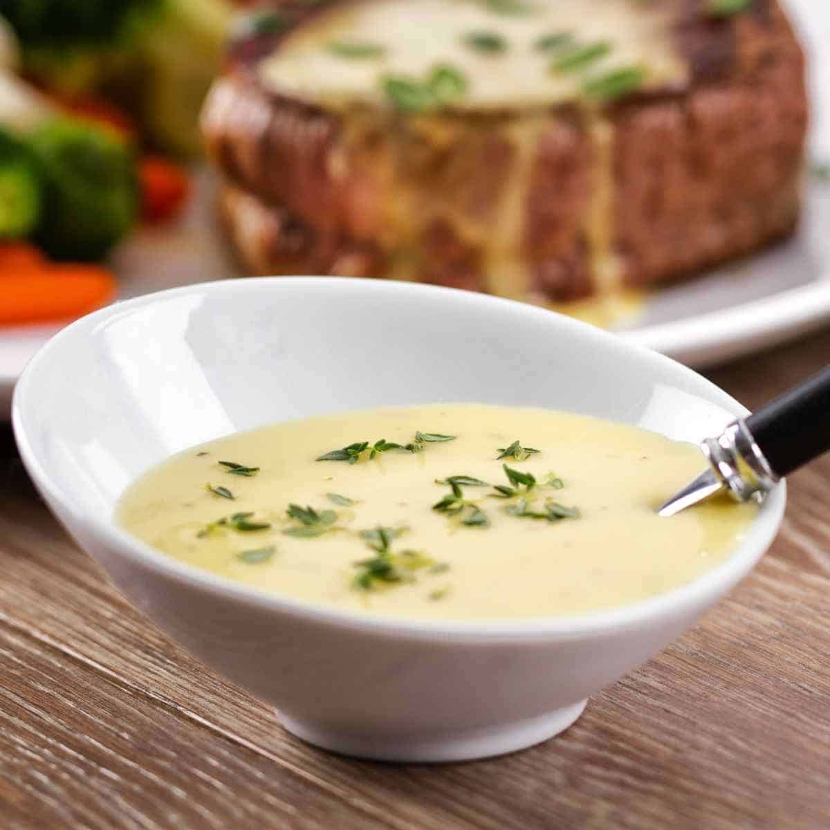 White bowl with cream colored sauce sprinkled with green leaves, a spoon sitting in bowl and a thick piece of beef with sauce on it set behind on a white plate with multi-colored vegetables all on a wood table.