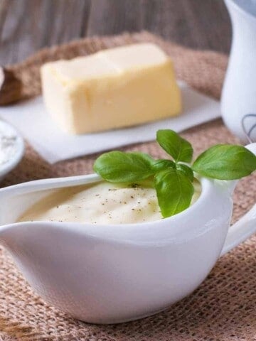 White sauce in a gravy boat with flour, butter, milk pitcher, and whisk on a burlap cloth.