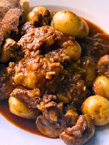 White bowl with brown beef chunks, mushrooms, onions, new potatoes in gravy with slice of bread.