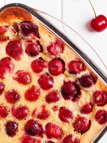 Clear square baking dish with cherries surrounded by a cream colored pudding on a white board background with one cherry beside baking dish.