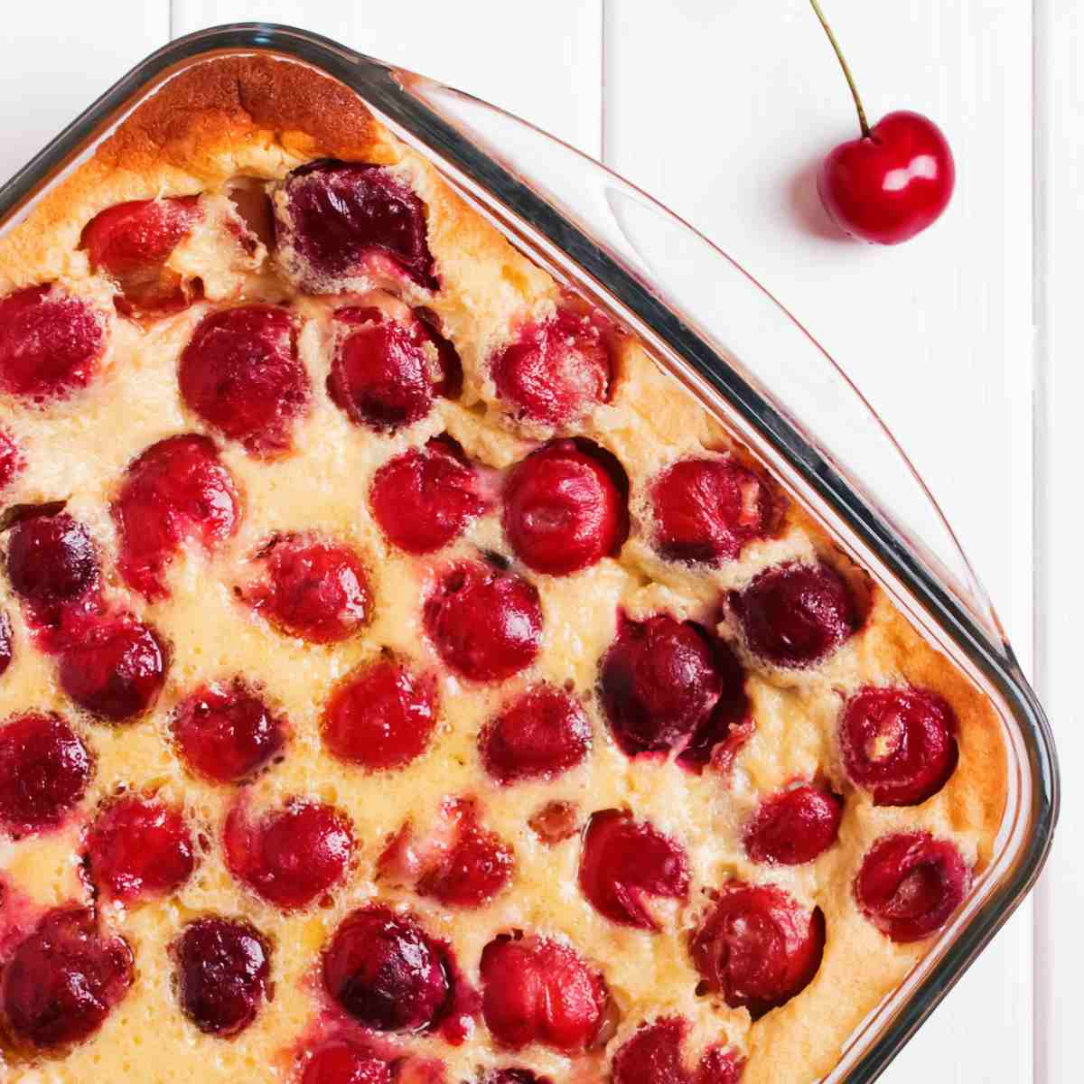 Clear square baking dish with cherries surrounded by a cream colored pudding on a white board background with one cherry beside baking dish.