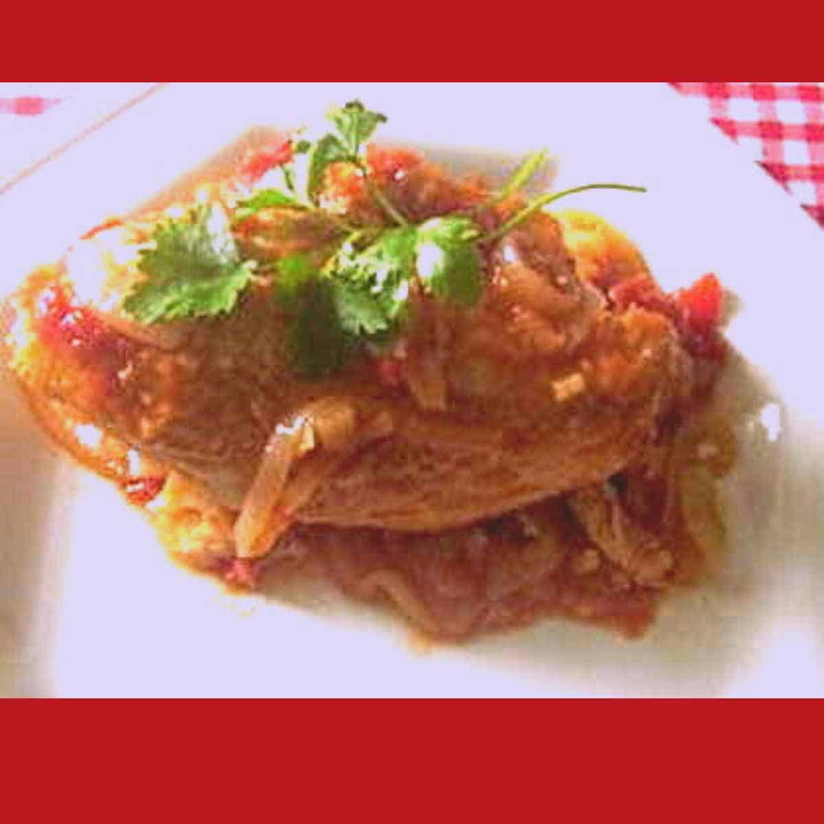 A chicken breast covered in red sauce with a sprig of parsley on top on a white plate with a red gingham table cloth.