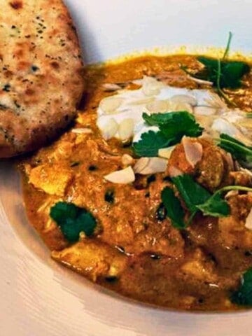 White bowl filled with a curry of flavorful chicken, onions, almonds, spices, and yogurt garnished with green leaves and spiced naan on the side.