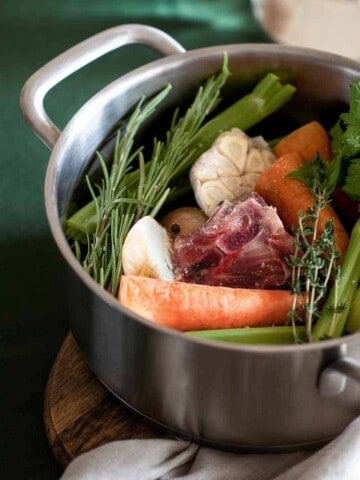 Steel pot filled with a red piece of meat, thyme, garlic, celery, carrots and onion on green surface with white cloth.
