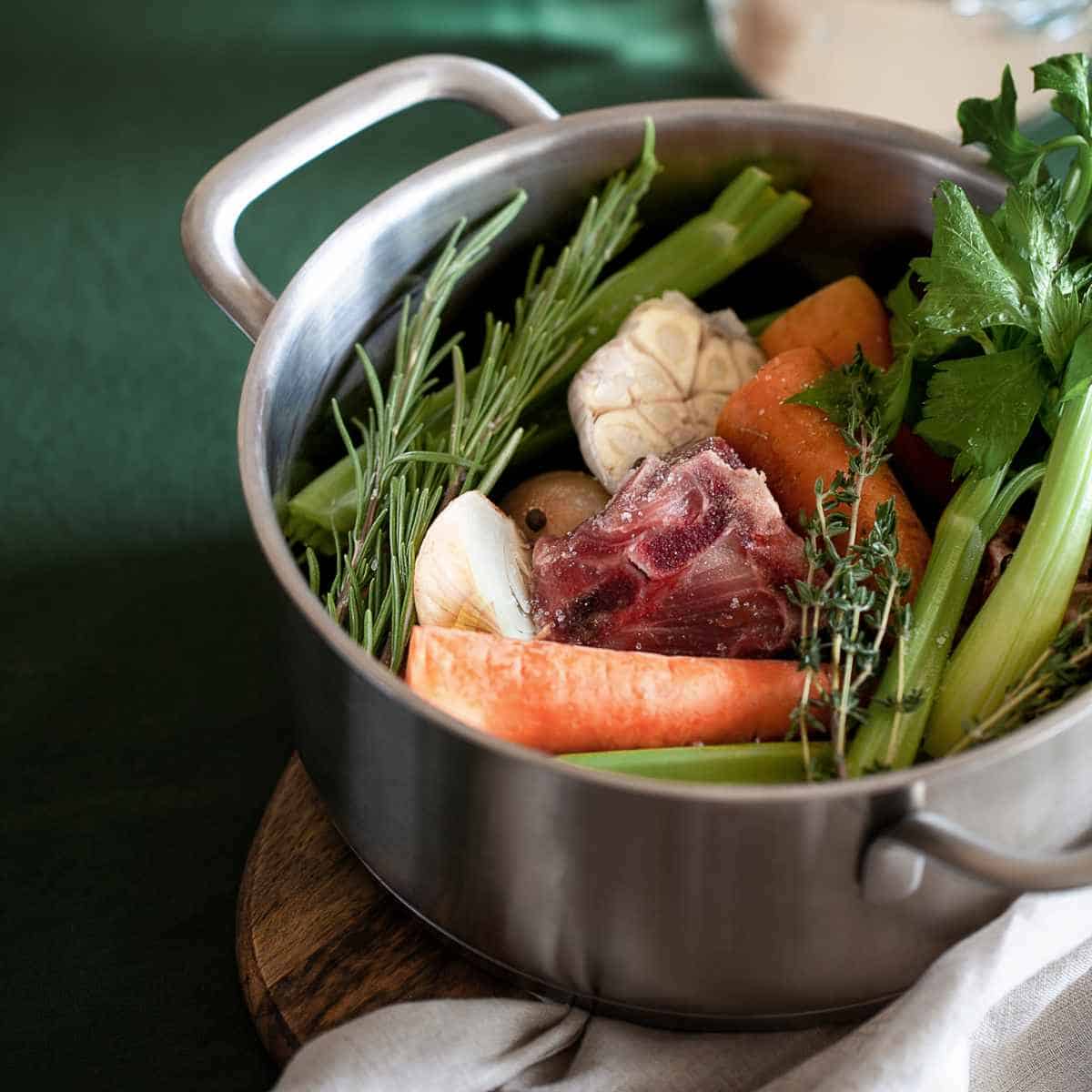 Steel pot filled with a red piece of meat, thyme, garlic, celery, carrots and onion on green surface with white cloth.