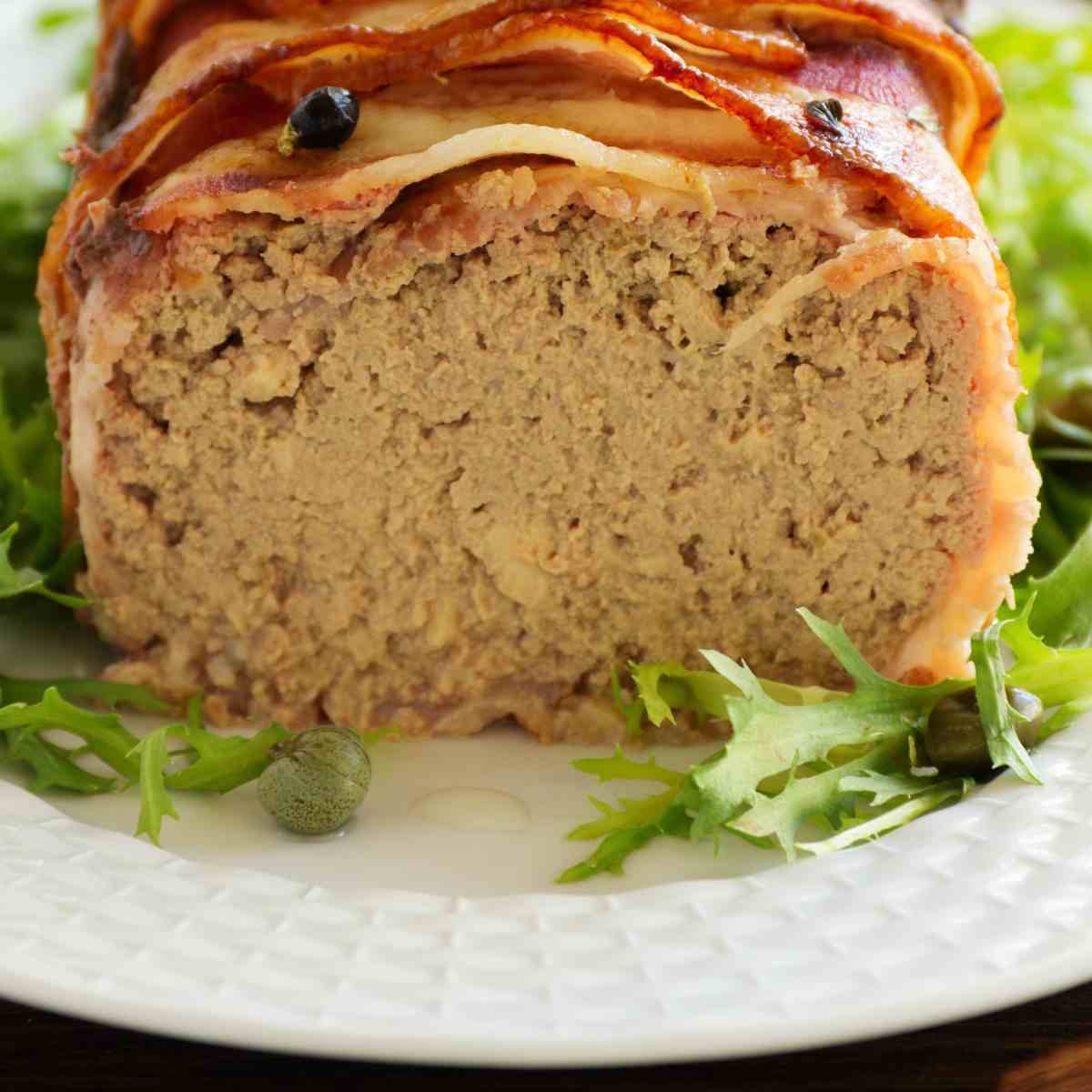 Light brown chicken liver paste wrapped in bacon on a white plate with a sprig of parsley.