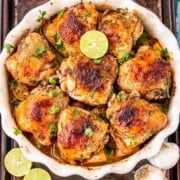 White round oven dish filled with well cooked chicken thighs with a half lemon and herbs on top with garlic bulbs and cut lemon on the side.
