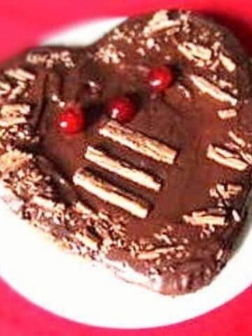 A heart shaped dark brown chocolate cake decorated with red candied cherries.