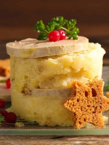 Stack of white mashed potatoes with two slices of light brown liver pate in-between and on top with garnishes of brown star shaped croutons, green parsley, and red berries.