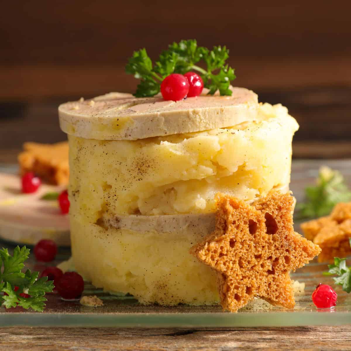 Stack of white mashed potatoes with two slices of light brown liver pate in-between and on top with garnishes of brown star shaped croutons, green parsley, and red berries.