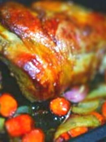 Large golden brown leg of lamb in roaster surrounded by green, pinkish, and orange vegetables.