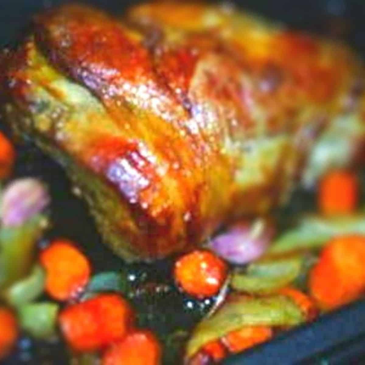 Large golden brown leg of lamb in roaster surrounded by green, pinkish, and orange vegetables.