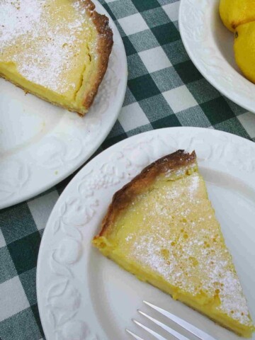 A slice of yellow lemon tart, sprinkled with white icing sugar with a brown crust.
