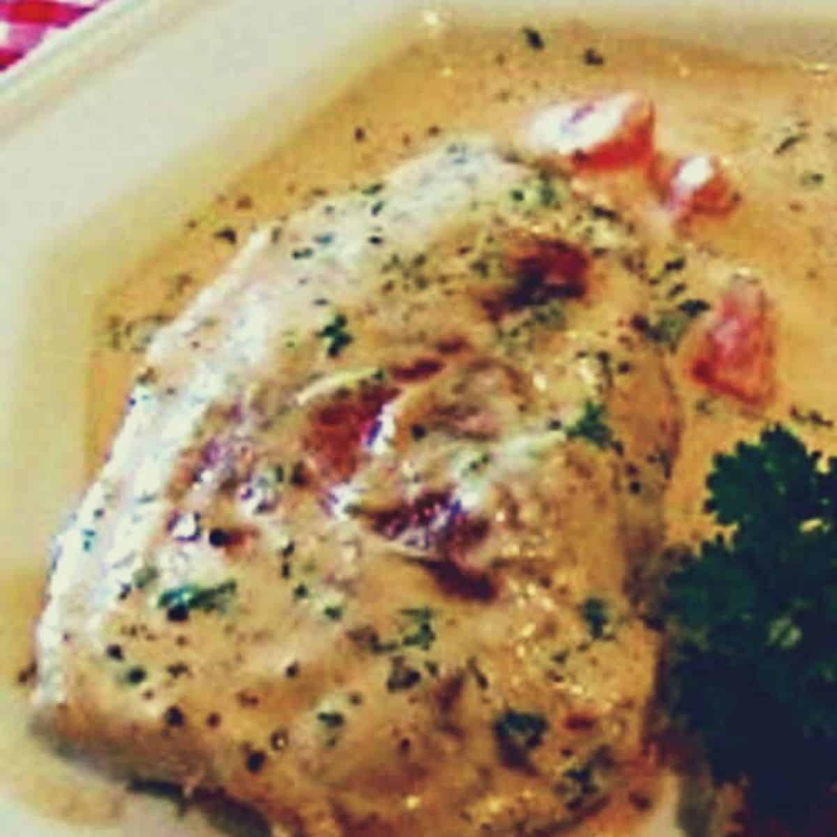 A white plate with a pork chop covered in white creamy sauce with pieces of tomato, with green parsley on the side.