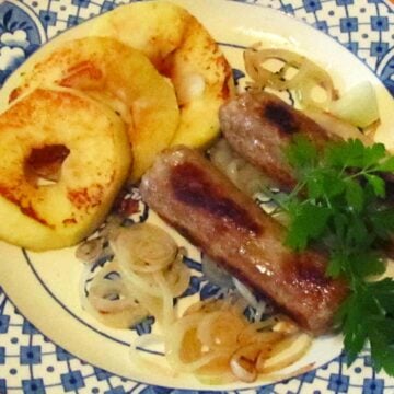 Two thick brown pork sausages on a blue patterned dinner plate with cooked onion and yellow sliced apples and green parsley garnish.