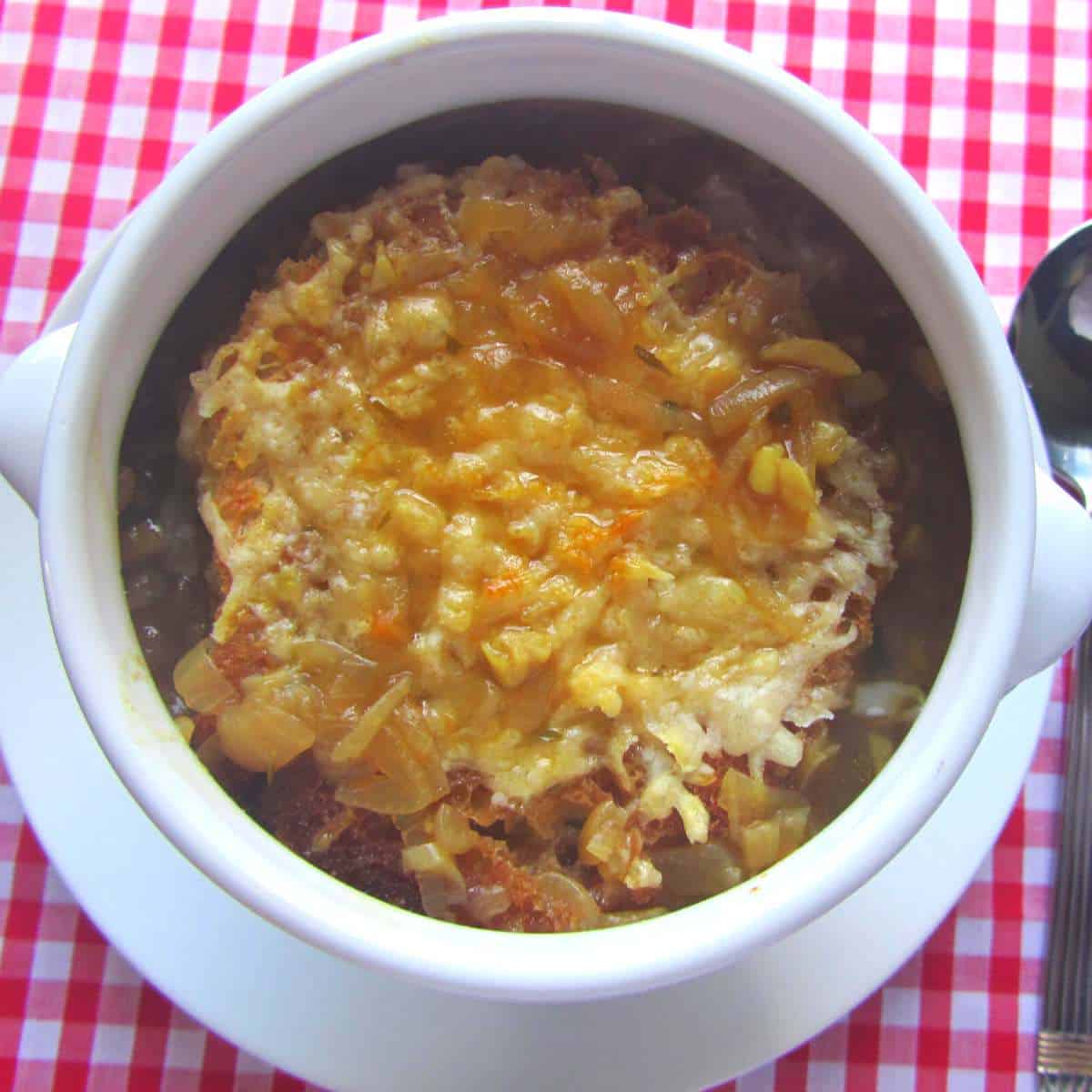 A white soup bowl on a red gingham table cloth filled with dark onion soup, topped with a slice of French bread covered in cheese.