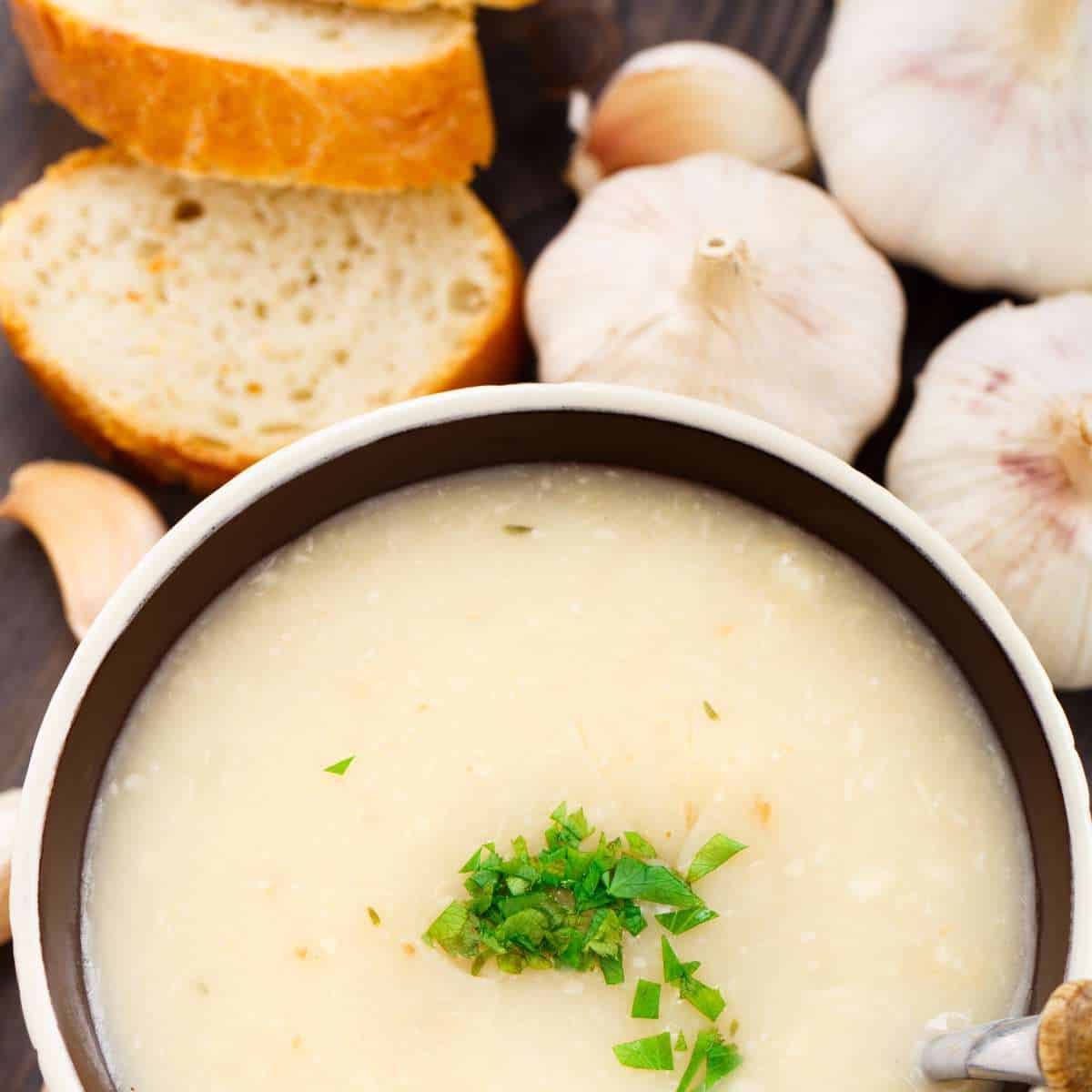 A white soup bowl with blue band inside filled with white creamy soup, sprinkled with green chives all beside sliced baguette and whole garlic cloves.
