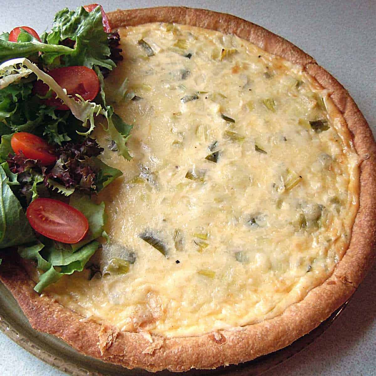 A round dish like a pizza but yellow in the center with green bits and light brown crust and a green lettuce and red tomato salad on one edge.
