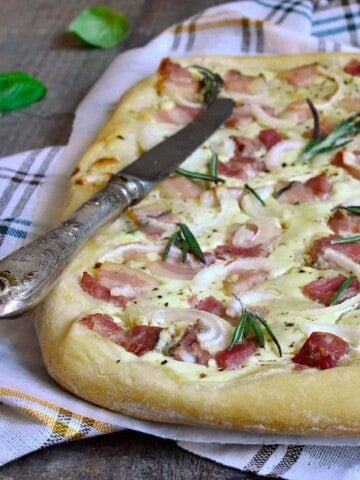 A pizza like food made with bread dough, cheese, bacon, and onions, on a kitchen towel and table knife.