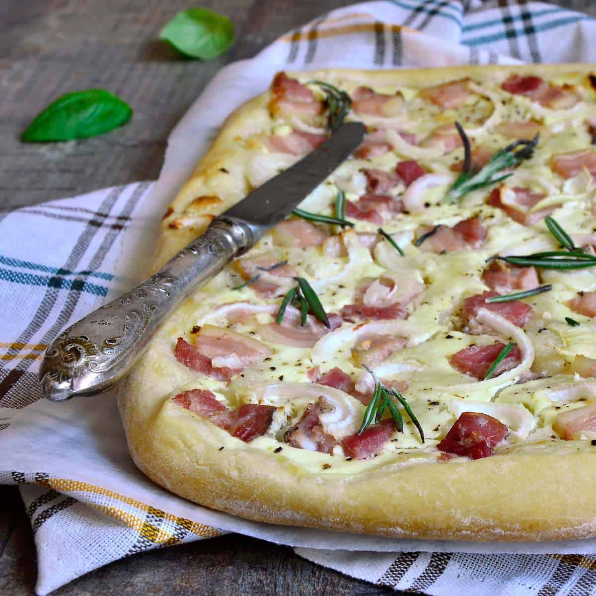 A pizza like food made with bread dough, cheese, bacon, and onions, on a kitchen towel and table knife.