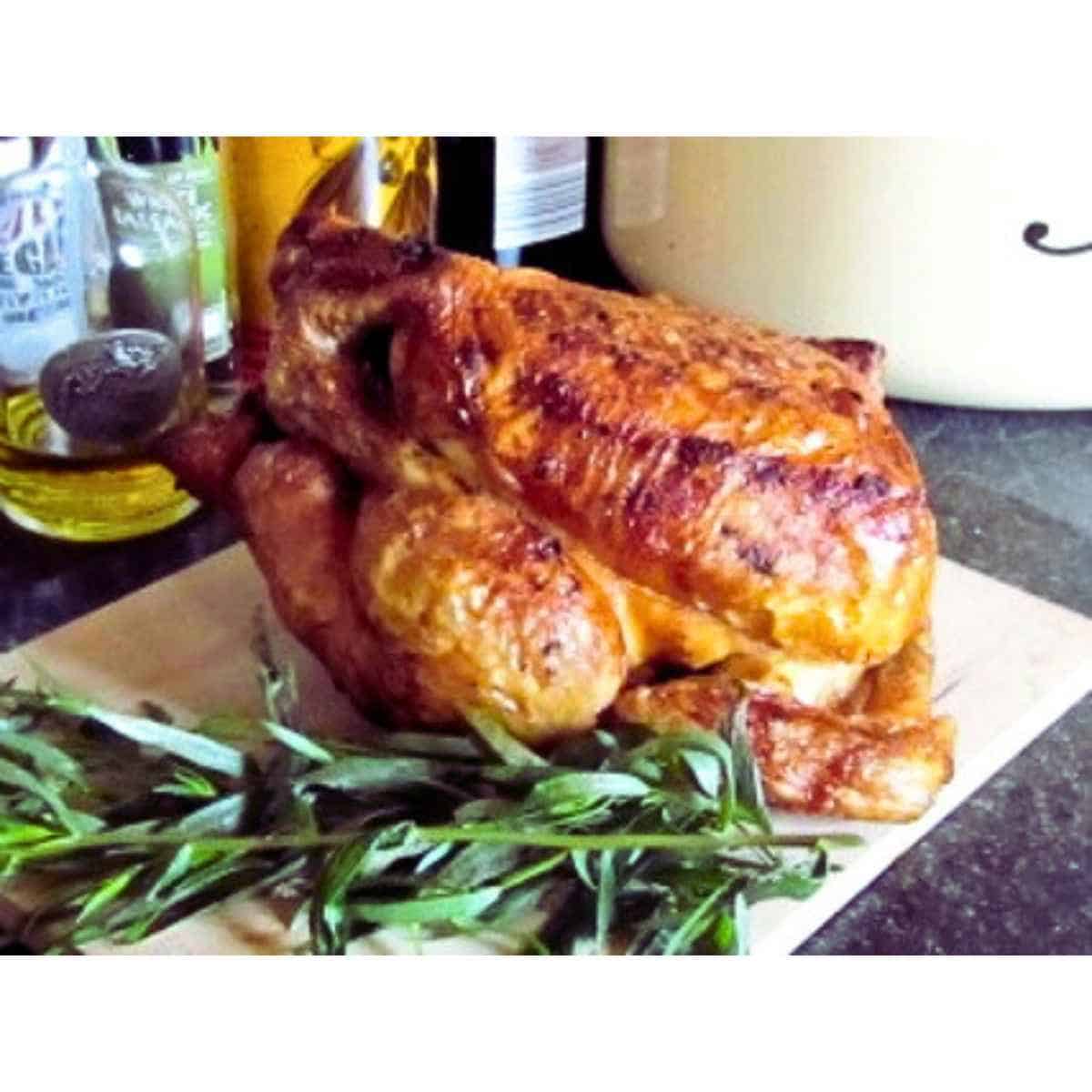 Roasted whole chicken with sprig of tarragon on a white cutting board.