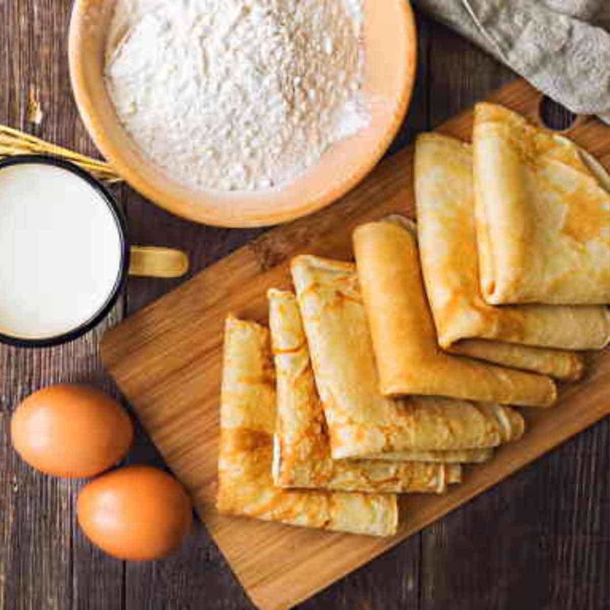 Six folded crepes on a wooden board surrounded with two eggs, salt and flour.