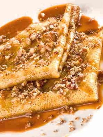 Two folded yellow crepes topped with brown caramel sauce and chopped beige pecans on white plate.