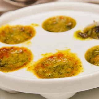 White dish with holes filled with butter, colored herbs, and snails.