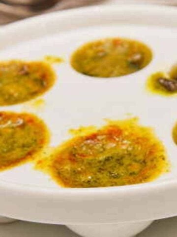 White dish with holes filled with butter, colored herbs, and snails.