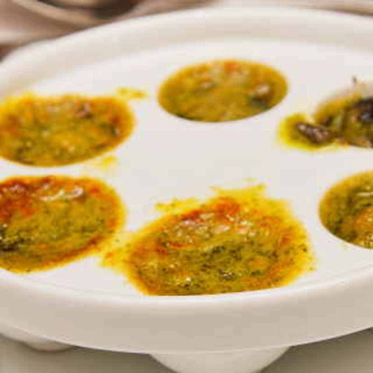 White dish with holes filled with butter, colored herbs, and snails.