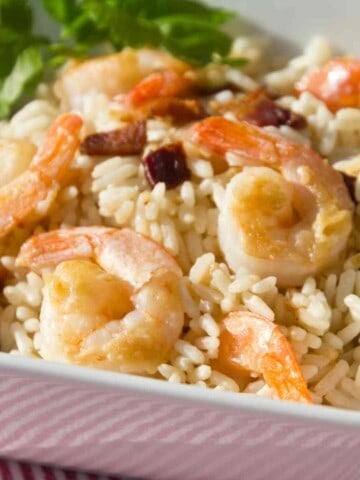 Square white bowl filled with rice, shrimp, and bacon with green leaf garnish.