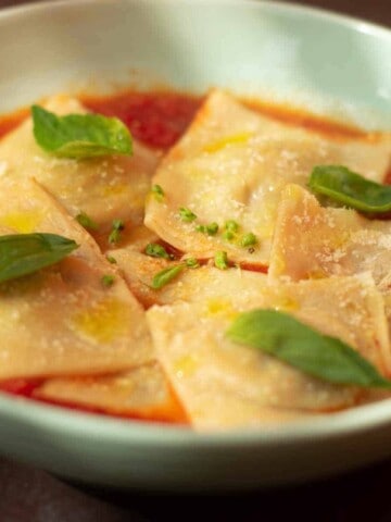 A white bowl with ravioli in tomato sauce with basil leaves.