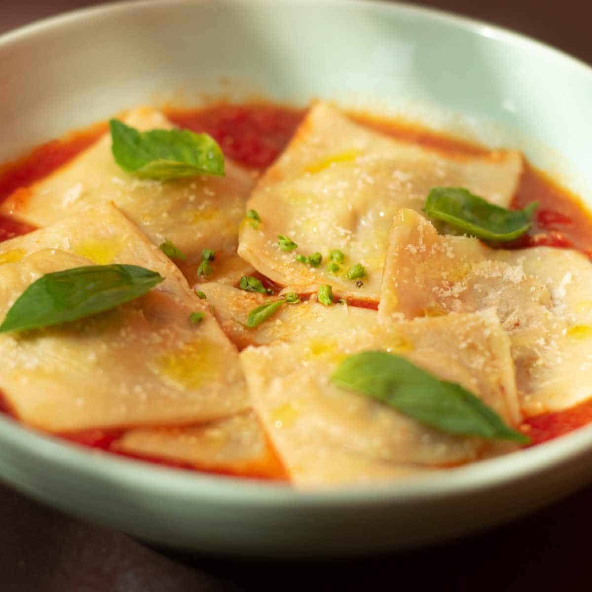 A white bowl with ravioli in tomato sauce with basil leaves.