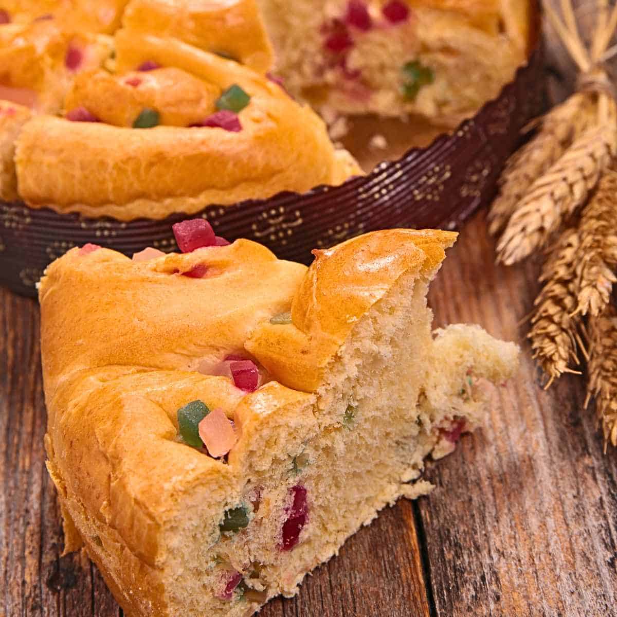 Round golden bread with red and green fruit pieces on a brown plate with a slice beside it and wheat sheafs on wood background.