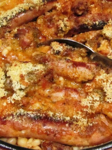 Large red cast-iron pot filled with sausage, beans, bacon, covered with bread crumbs.