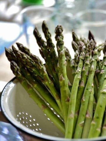 Multiple green spears of asparagus standing up in a white metal colander, over a sink with 2 glasses in the background.