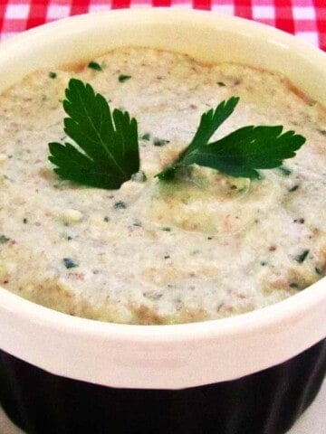 White and black bowl filled with creamy, pate with bits of mushroom garnished with parsley on a red gingham table cloth.