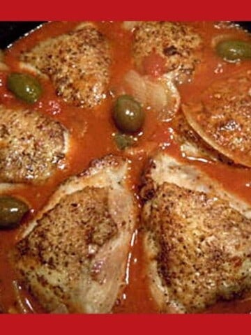 Black cast iron frying pan with chicken thighs in tomato sauce wth olives.