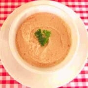 Pink spread with parsley garnish in a white bowl on top of a white plate on a red gingham table cloth.
