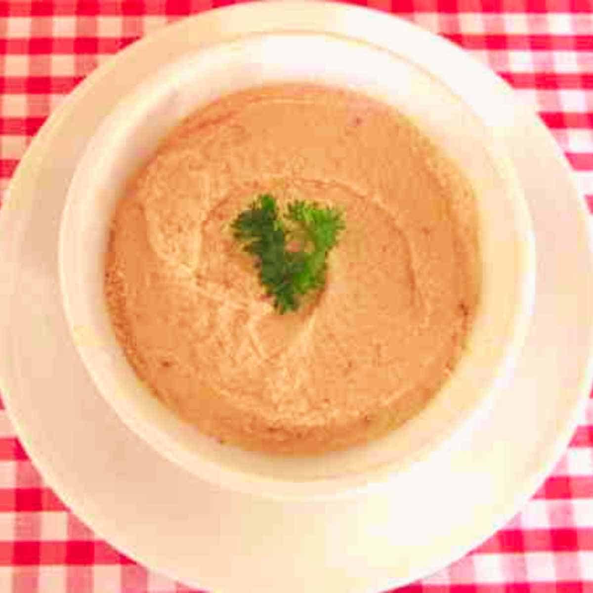 Pink spread with parsley garnish in a white bowl on top of a white plate on a red gingham table cloth.