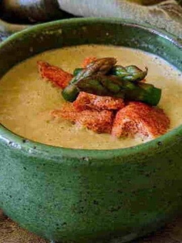 Green earthen bowl on cloth full of green soup with chopped brown bread and green asparagus heads in center of soup.