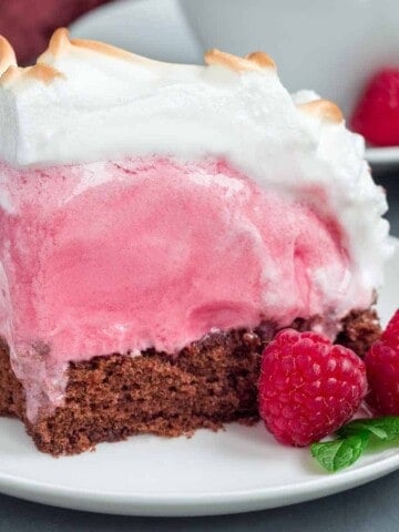 Dark cake layered with pink ice cream and covered with white meringue on a white plate with two raspberries.