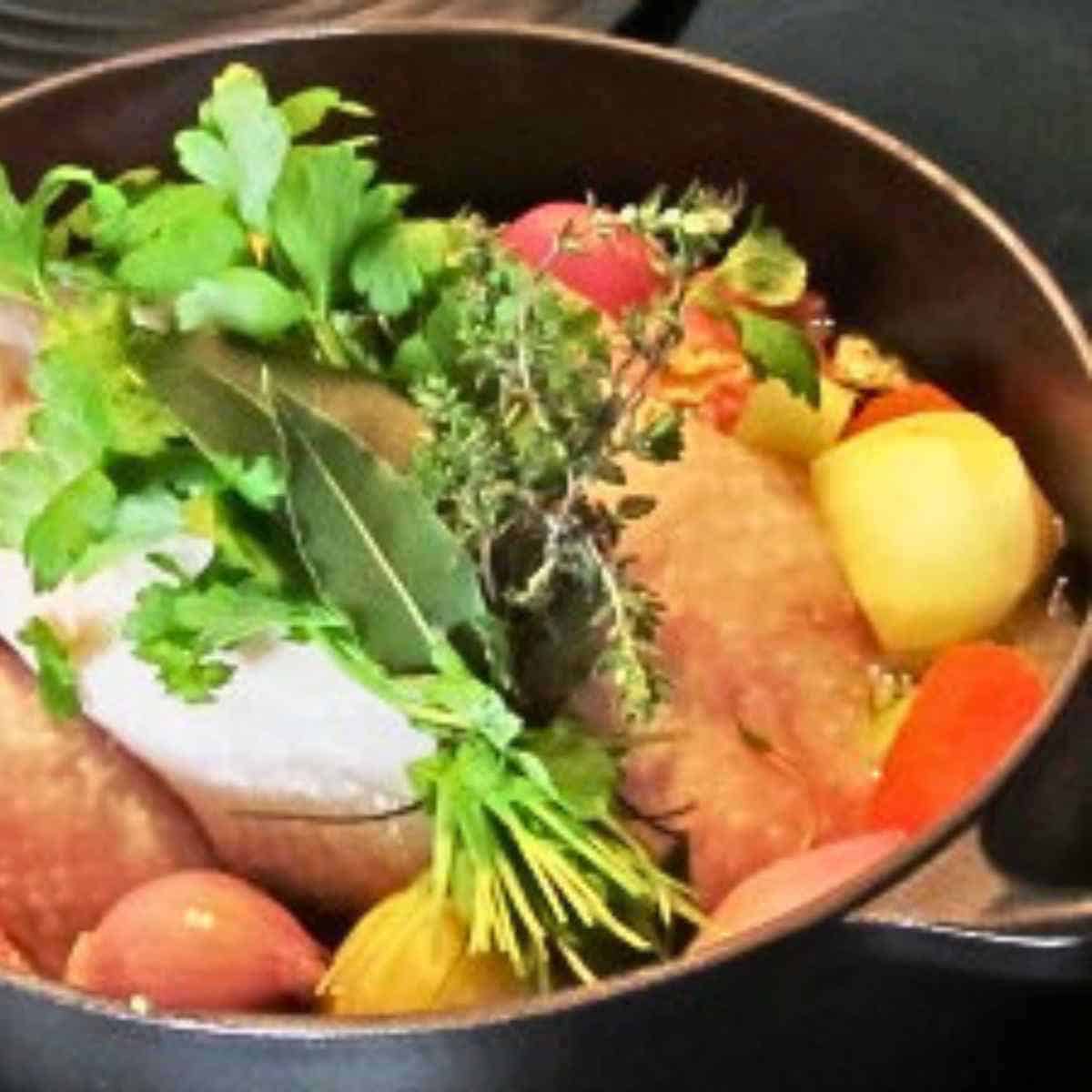 Large black pot full of chicken pieces some white, some biege with skin on, yellow potatoes, orange carrots, onions with brown skin on, an green herbs.