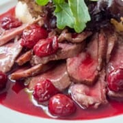 Sliced duck breast meat covered in cherries and red sauce on a white plate with salad on the side.